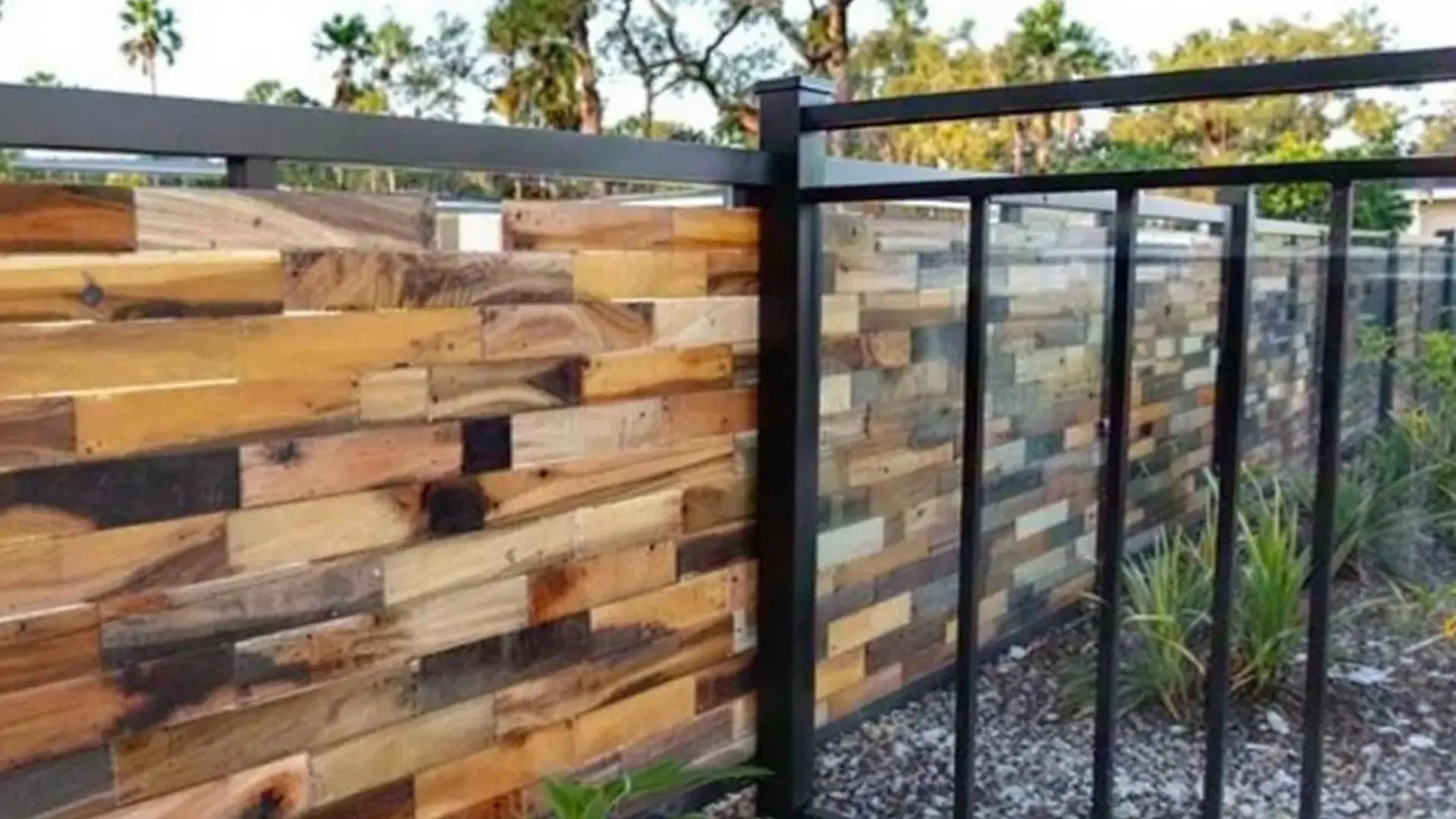 A modern fence made of horizontal wooden planks in varying natural tones, framed with black metal and glass panels. The fence is set along a landscaped area with small plants and gravel, with trees visible in the background.