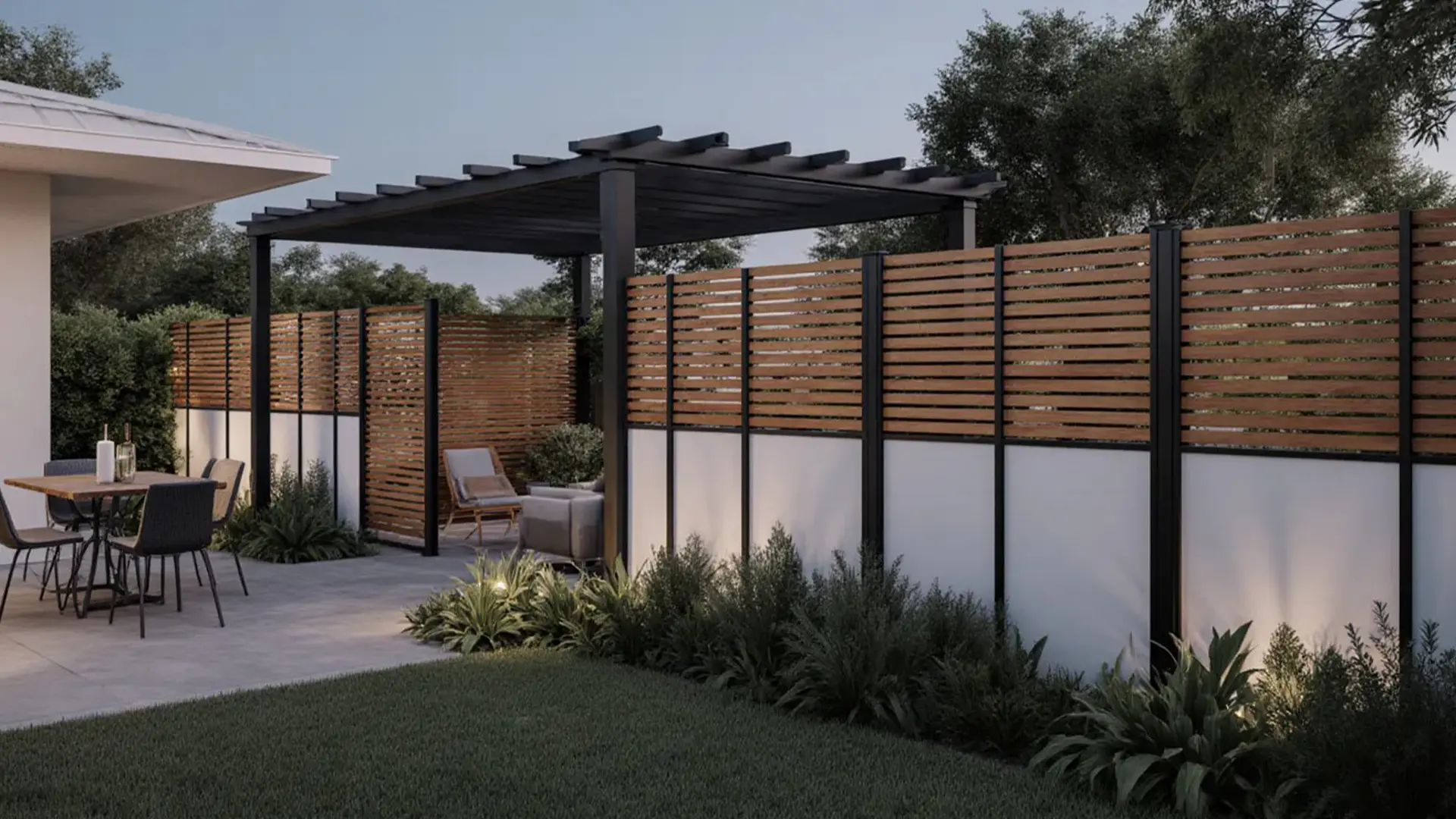 A landscaped backyard patio features a black pergola with horizontal beams positioned beside a modern privacy fence made of horizontal wood slats framed in dark metal, with solid light-colored panels along the lower section. An outdoor dining table and chairs sit on a paved patio area near the house, while low shrubs and plants line the base of the fence. The scene is set at dusk, with soft, even lighting and mature trees visible in the background.